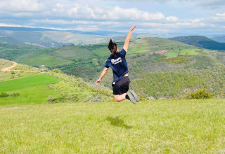 Person springt auf einer Wiese mit Hügeln im Hintergrund bei Flower Camping Le Pont du Tarn, Occitanie, Frankreich.