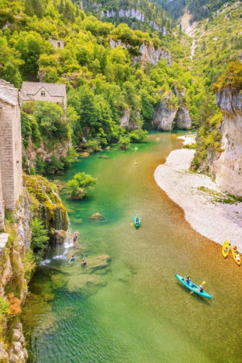 Kayakers paddle on a vibrant green river by cliffs and old buildings near Florac-Trois-Rivières, Occitanie, France.
