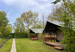 Safari-telte på Camping 'de Val' i Holland, omgivet af grønne træer og en græsplæne under blå himmel.
