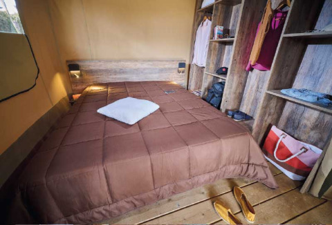 Innenansicht einer Safari-Lodge im Flower Camping Le Pont du Tarn mit Doppelbett und Holzregalen.