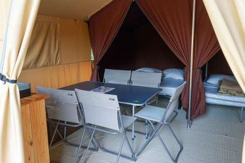 Intérieur d'une tente safari avec table à manger, chaises pliantes et deux lits sur un sol en moquette.