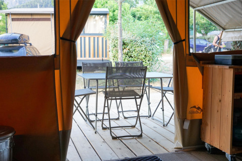 Vue depuis l’intérieur d’une tente safari sur une terrasse avec table, chaises et verdure.