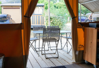 Vue depuis l’intérieur d’une tente safari sur une terrasse avec table, chaises et verdure.