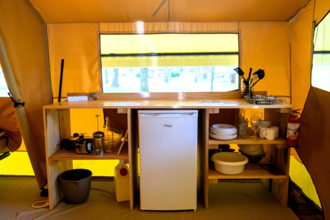 Zona de cocina dentro de una tienda safari con frigorífico pequeño, utensilios, vajilla y extintor.