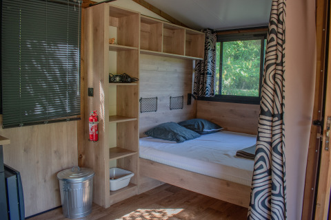 Interior de una tienda safari con cama doble, estanterías de madera, ventanas grandes y cortinas decoradas.