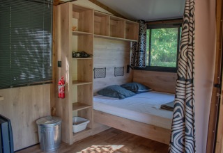 Interior de una tienda safari con cama doble, estanterías de madera, ventanas grandes y cortinas decoradas.