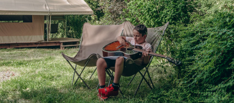 Niño toca la guitarra sentado en una silla de camping en Onlycamp Camping de Besançon, rodeado de verde.