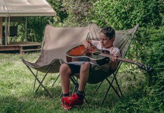 Boy playing guitar while relaxing in a camping chair at Onlycamp Camping de Besançon, surrounded by nature.