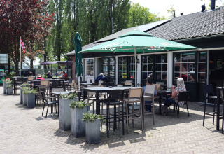 Outdoor lodge patio with tables, chairs, parasols, and guests enjoying a sunny day among green trees.