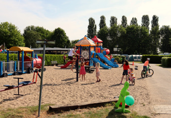Children play on a sandy playground with slides and bicycles next to a cozy lodge on a sunny day.