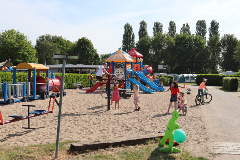 Children play on a sandy playground with slides and bicycles next to a cozy lodge on a sunny day.