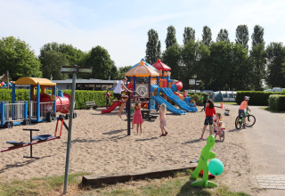 Children play on a sandy playground with slides and bicycles next to a cozy lodge on a sunny day.