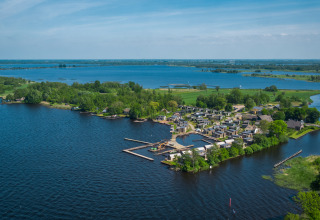 Vista aerea di un glamping sul lago con bungalow moderni, pontili in legno e vegetazione rigogliosa.