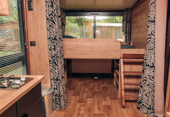 Tiny house interior with wood flooring, lofted bed, patterned curtains, and large window at the rear.