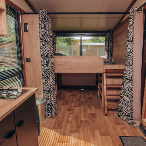 Innenraum eines Tiny Houses mit Holzfußboden, Hochbett, Vorhängen und großem Fenster im Hintergrund.