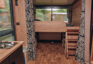 Innenraum eines Tiny Houses mit Holzfußboden, Hochbett, Vorhängen und großem Fenster im Hintergrund.