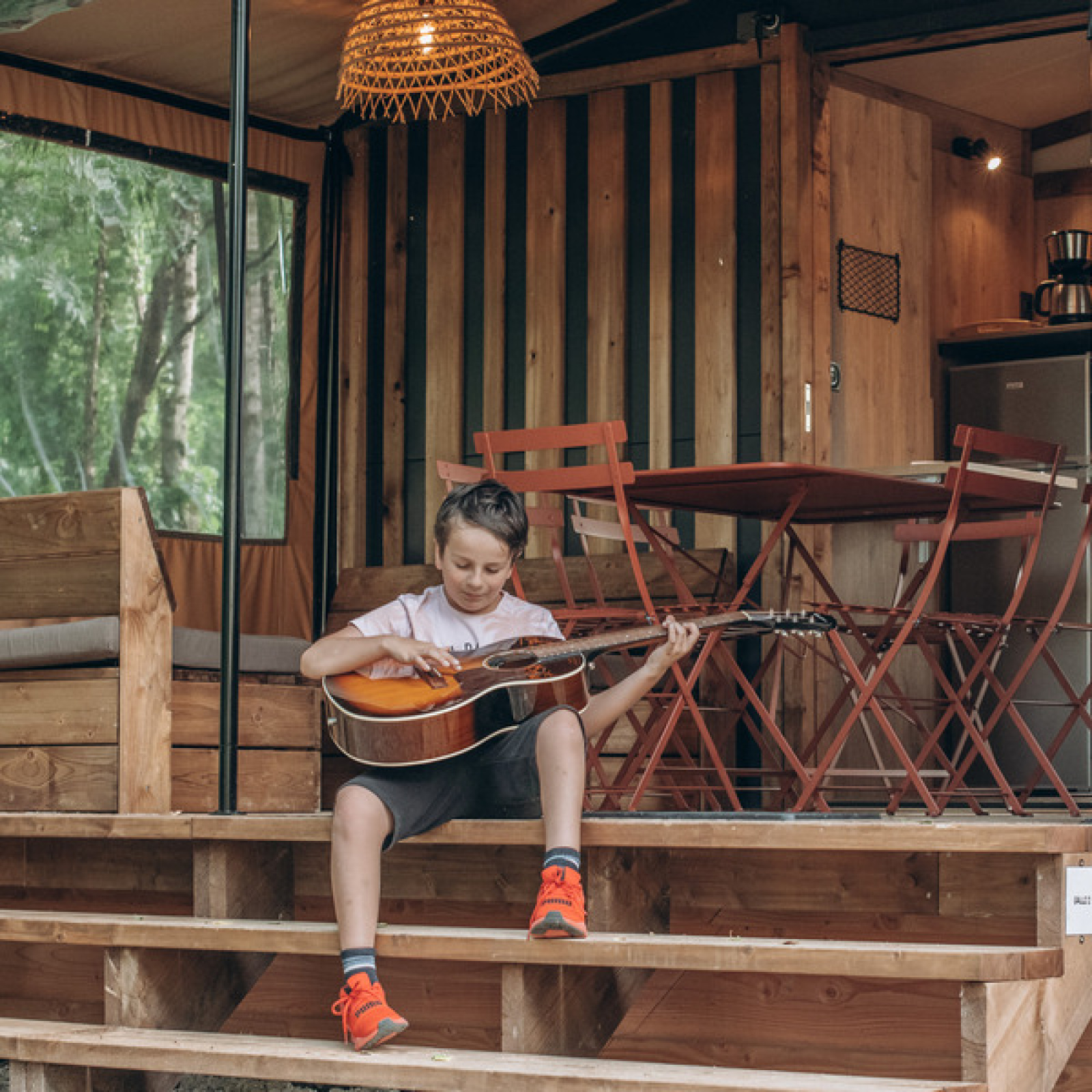 Jongen speelt gitaar op de trap bij het Chalet Galli tiny house op Onlycamp Camping de Besançon, Frankrijk.