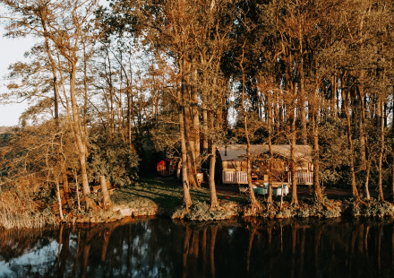 Safaritent Glamphuys Ultra Wellness ligt beschut tussen bomen aan een rustig meer, omringd door natuur.