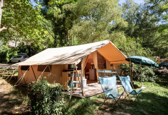 A Ponza safari tent campsite with open awning, outdoor chairs, and umbrella set in a green forest area.
