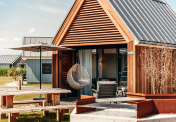 Tiny wooden house with a hanging chair, umbrella, picnic table, and fire pit in front of Tiny House 2.