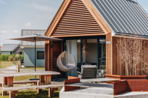 Lille hytte i træ med gyngestol, parasol, picnicbord og udendørs bålplads foran Tiny House 2.