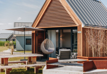 Pequeña casa de madera con silla colgante, sombrilla, mesa de picnic y fogata frente a Tiny House 2.