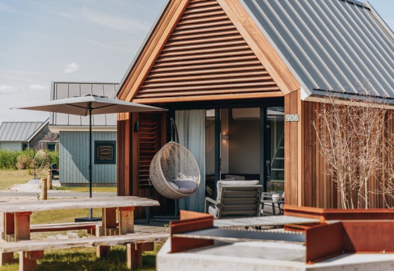Tiny wooden house with a hanging chair, umbrella, picnic table, and fire pit in front of Tiny House 2.