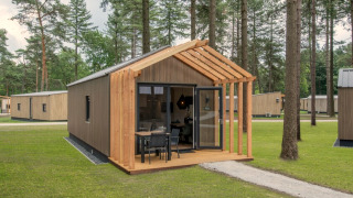 Tiny House 2, een klein houten huisje met open veranda, tuintafel en stoelen in een groene bosomgeving.