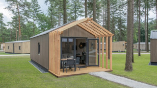 Lille træhus kaldet Tiny House 2 i en skov, med terrasse og havemøbler foran indgangen.