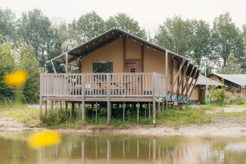 Tente safari de luxe au bord de l'eau avec terrasse en bois sur pilotis, entourée de forêt verdoyante.