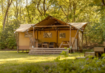 Safari-Zelt im Wald mit einer Holzterrasse und Sitzgelegenheiten im Freien, umgeben von grünen Bäumen.