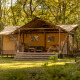 Safari tent with wooden deck and outdoor seating in a forest setting, surrounded by tall green trees.