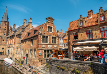 Photo of a scenic canal, historic brick buildings, and outdoor cafes with people on a sunny day in Europe.