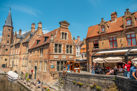 Photo of a scenic canal, historic brick buildings, and outdoor cafes with people on a sunny day in Europe.