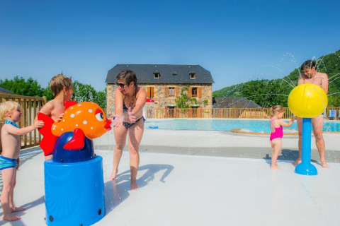 Family with kids playing by water toys at a pool in a holiday park, stone house, and hills in the background.
