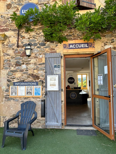 Ingresso della reception di Seasonova Camping Les Rives d'Olt et Aubrac con bacheca e sedia nera fuori.