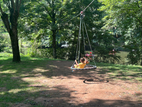 Un bambino si rilassa su un'altalena rotonda sotto alberi alti vicino a un fiume a Seasonova Camping Les Rives d'Olt et Aubrac, Francia.