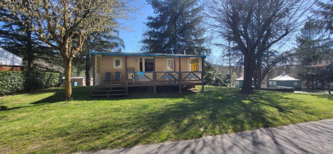 Mobile Home Eco with a wooden porch and chairs in a sunny grassy area with trees in the background.