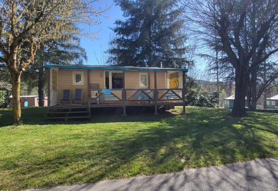 Mobile Home Eco with a wooden porch and chairs in a sunny grassy area with trees in the background.