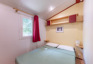 Gemütliches Schlafzimmer mit Doppelbett, Fenster und roten Vorhängen in Mobile Home Eco, Val de Coise, Frankreich.