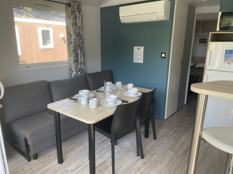 Zona pranzo con divano grigio e tavolo apparecchiato per quattro nel mobilhome Comfort+ al Seasonova Camping Le Val de Coise, Francia.