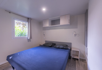 Dormitorio de casa móvil premium con cama grande, ropa de cama azul, ventana y almacenamiento moderno en Francia.