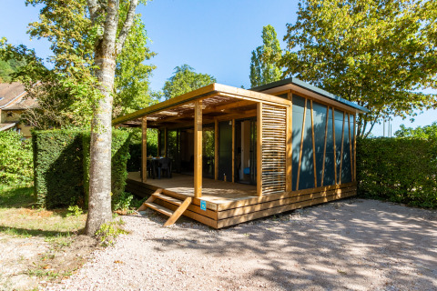 Moderne glampingaccommodatie met houten veranda, omringd door bomen en veel groen op een zonnige dag.