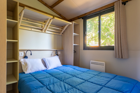 Soveværelse med dobbeltseng, blå dyne, åbne hylder og vindue i Cottage Comfort på Camping Paradis Le Giessen.