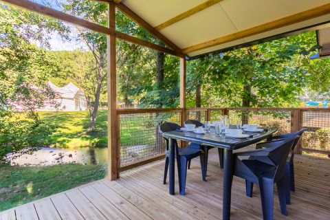 Terraza de madera con mesa de comedor en Cottage Comfort, Camping Paradis Le Giessen, Francia, junto a un arroyo.