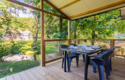 Terraza de madera con mesa de comedor en Cottage Comfort, Camping Paradis Le Giessen, Francia, junto a un arroyo.