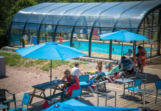 Udendørs pool med glastag og gæster, der slapper af under blå parasoller på Camping Paradis Les Chanterelles.