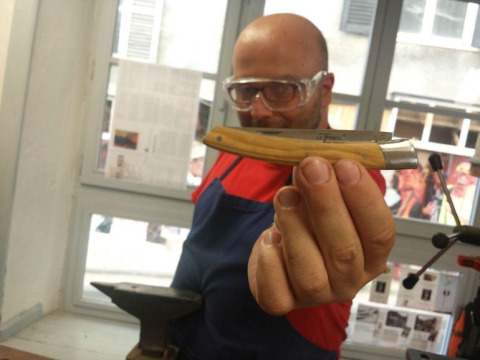 Hombre con gafas de seguridad y delantal muestra una navaja plegable en un taller junto a una ventana.