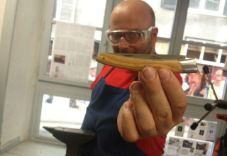 Hombre con gafas de seguridad y delantal muestra una navaja plegable en un taller junto a una ventana.