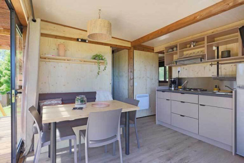 Interior luminoso de una cabaña con mesa de comedor, sillas, cocina y ventanales con vistas a la naturaleza.